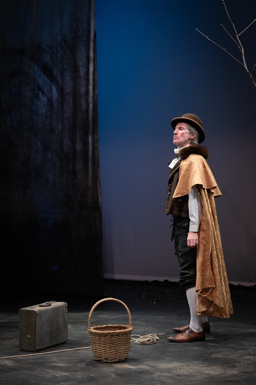 Pozzo in his cape and hat gazes upward, suitcase and basket at his feet