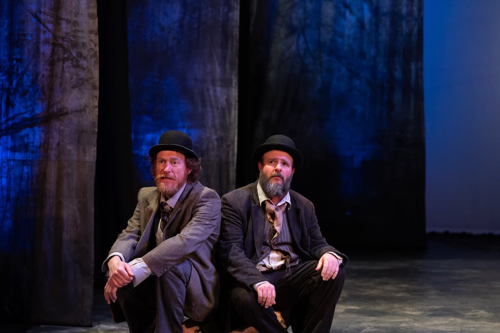 Vladimir and Estragon crouch side by side against a blue-lit backdrop